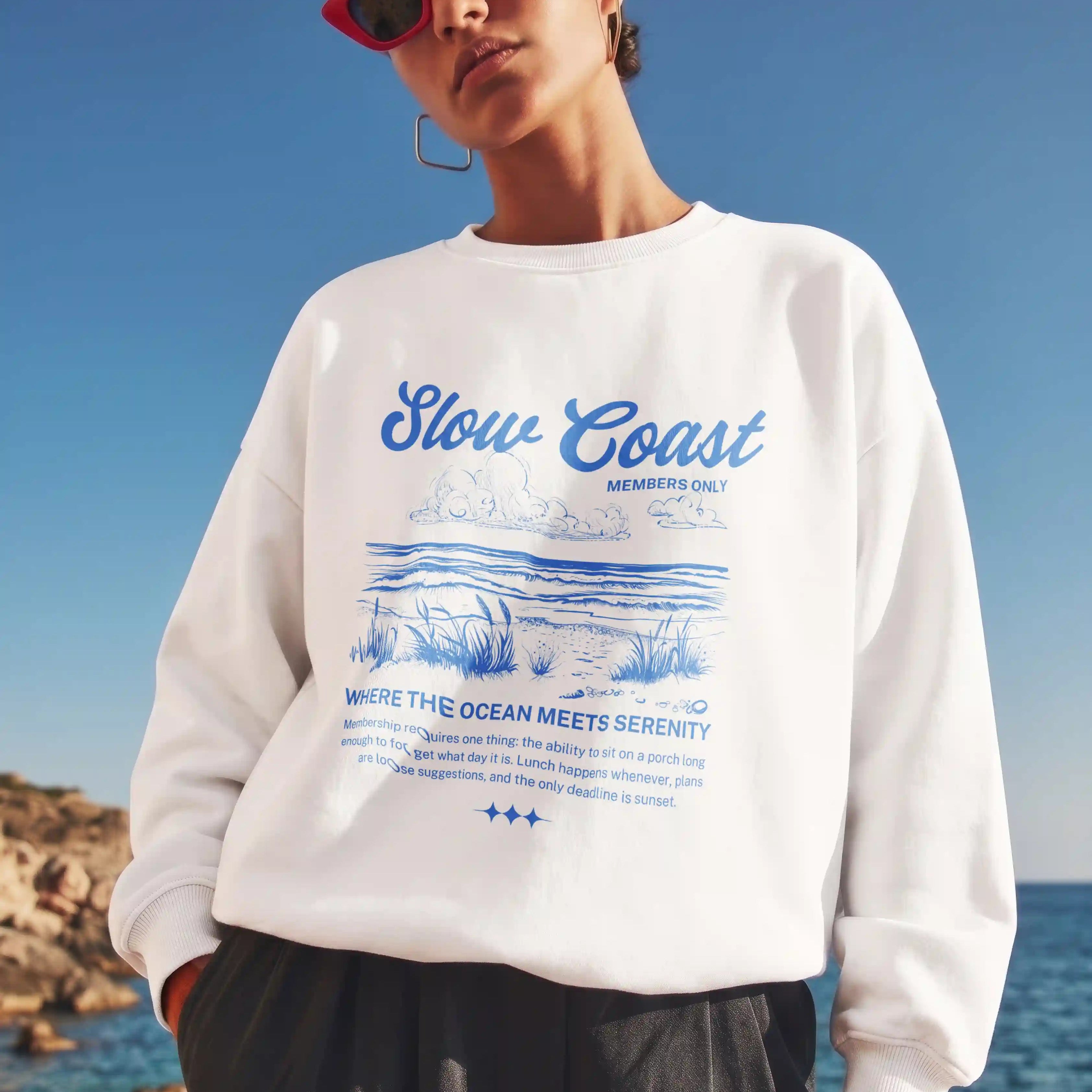 Person wearing a white sweatshirt with 'Slow Coast' design by the ocean