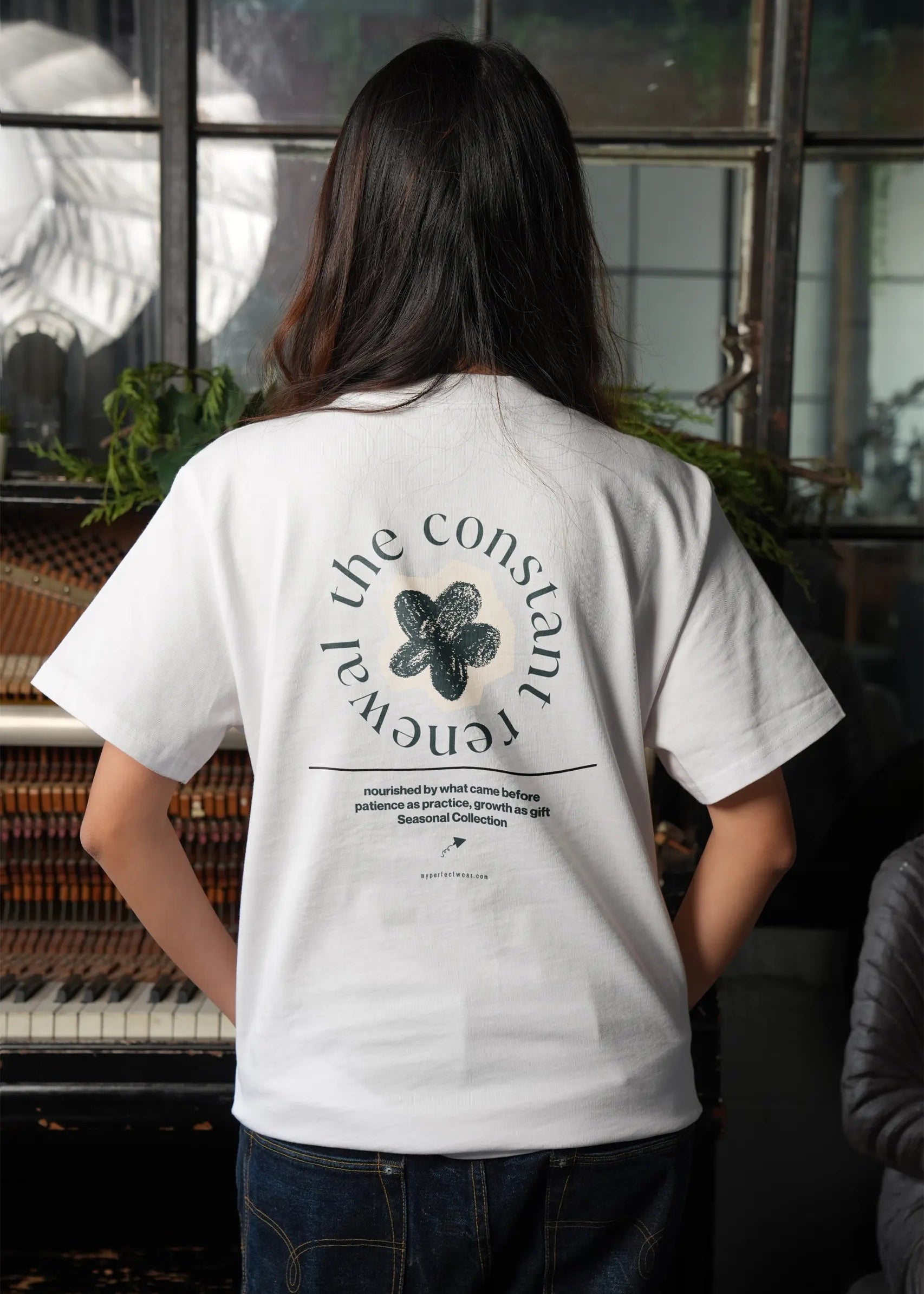 Person wearing a white t-shirt with a design and text on the back, standing in a room with a piano and plants.