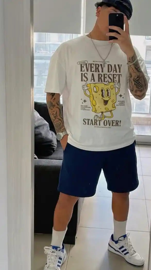Person wearing a white t-shirt with a graphic design and text, standing in a room with large windows.