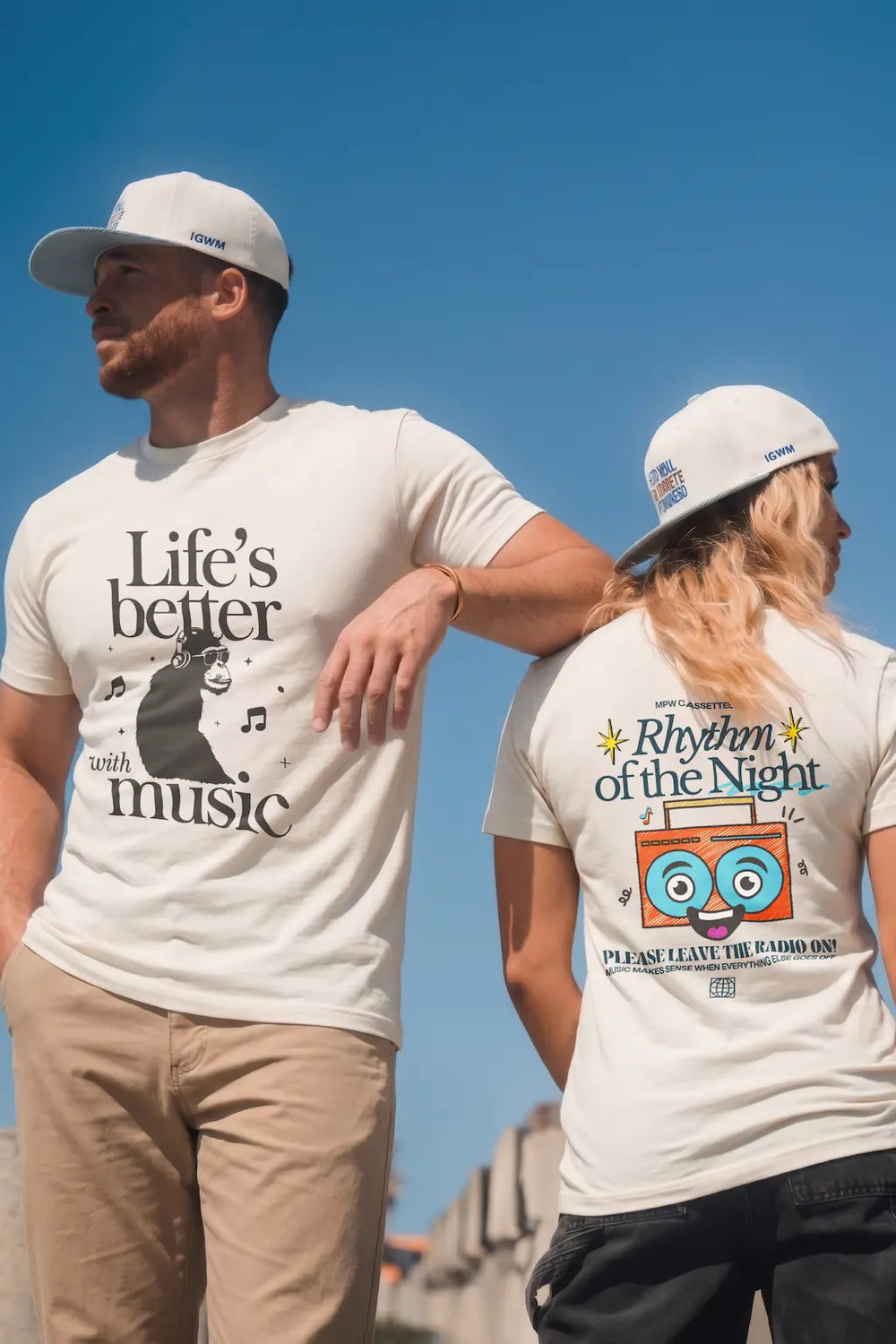 Man in white graphic tee reading ‘Life's better with music’ and women wearing white graphic tee reading 'Rhythm of the Night'  standing under blue sky